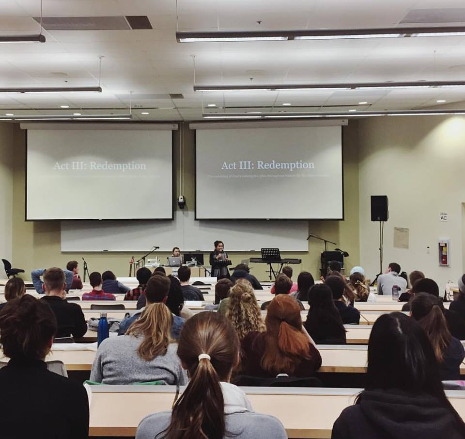 A woman speaks to a lecture hall full of young adults. The screen at the front says Act III: Redemption.