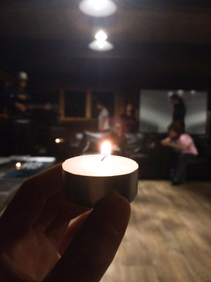 Picture of a hand holding a lit tea candle. There are people out of focus in the background. 